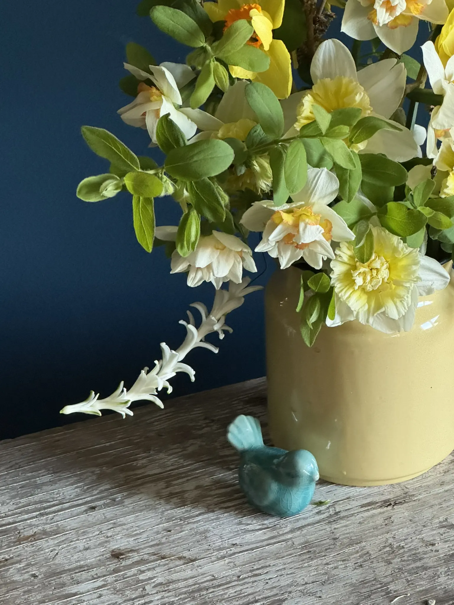 A yellow pot with white flowers and a small blue bird figurine on a wooden surface.