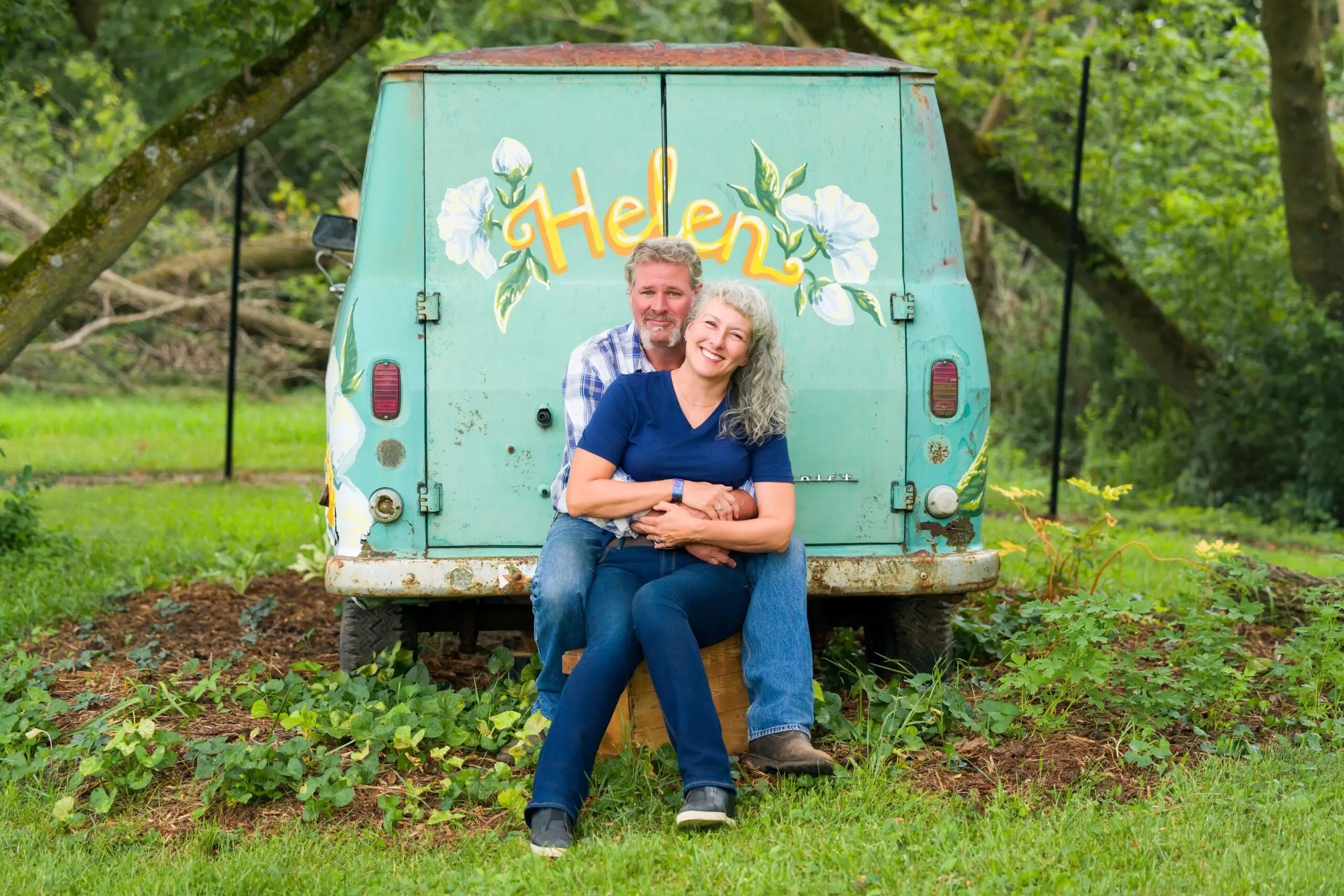 A happy couple embraces sitting in front of a vintage green van in a lush outdoor setting.