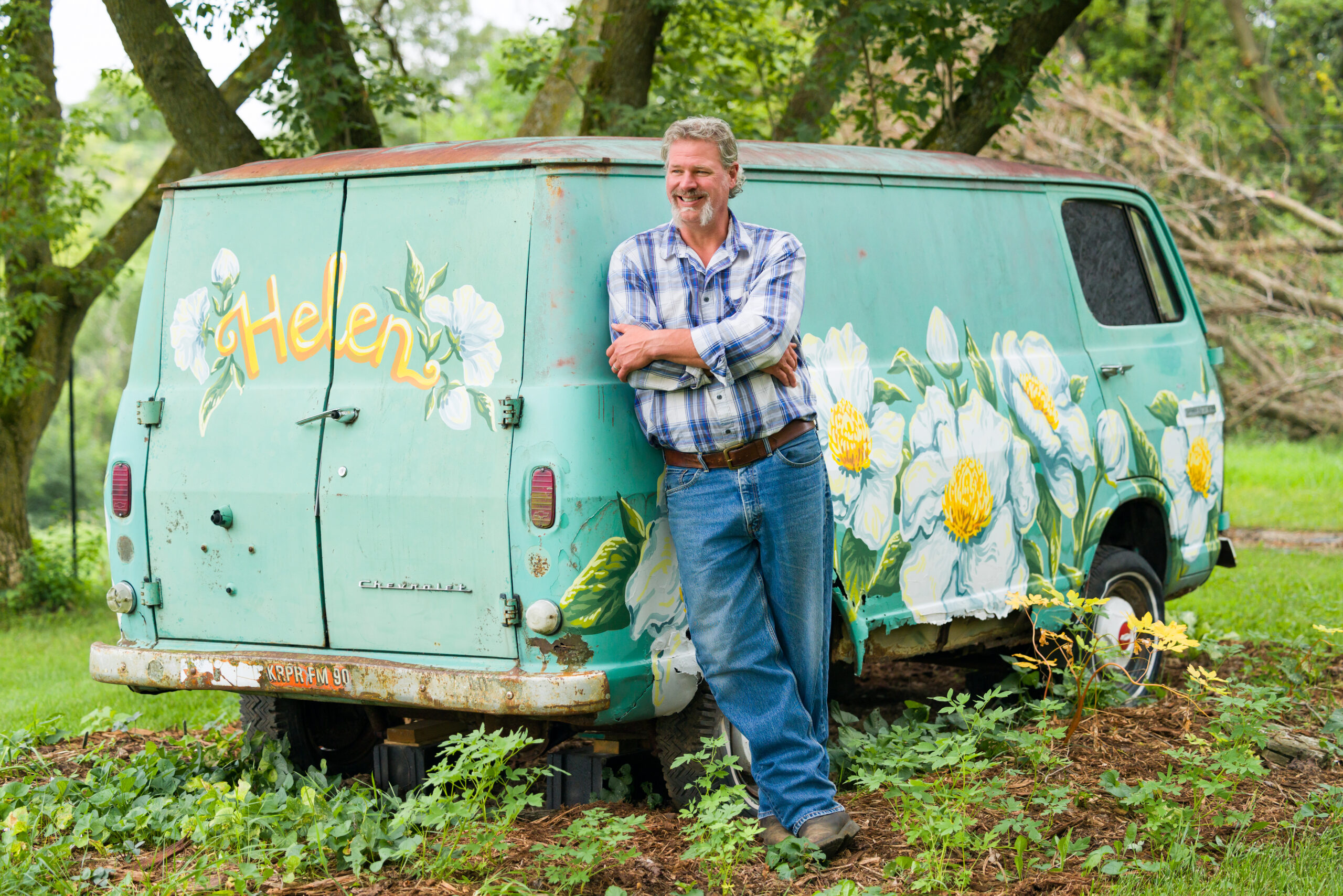 Man leaning against a vintage van painted with daisies in a lush garden.