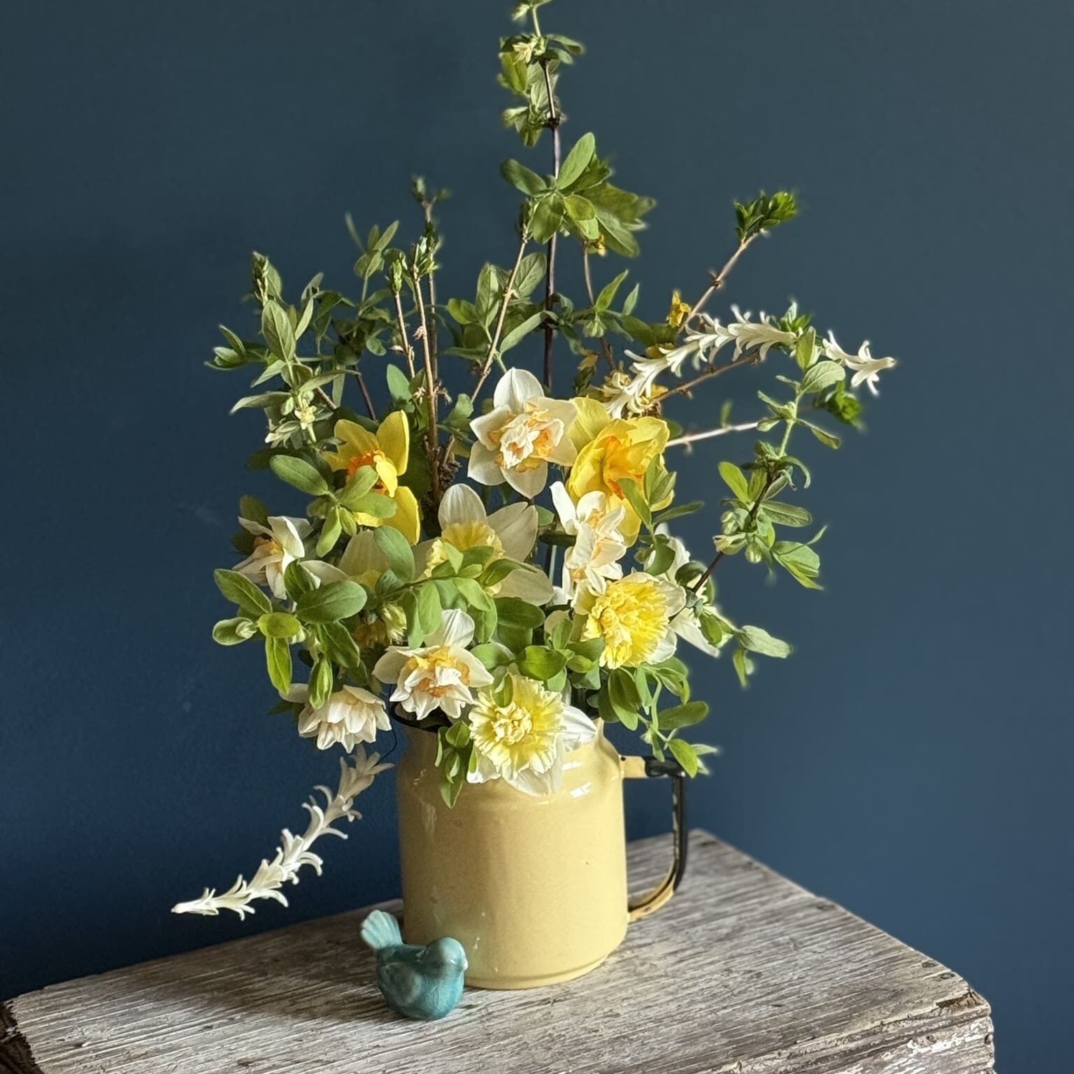 A yellow vase with fresh yellow and white flowers against a dark blue wall.